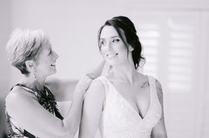 Black and white photo of a bride in a lace gown being helped by an older woman near a bright window.