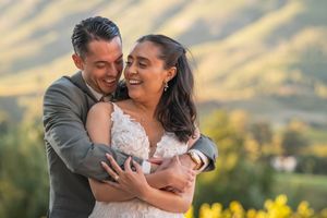A joyful couple embraces outdoors in wedding attire with lush green hills in the background.