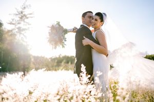 Bride and groom embrace in sunlit field of wildflowers, bride holding bouquet in elegant white gown and veil.