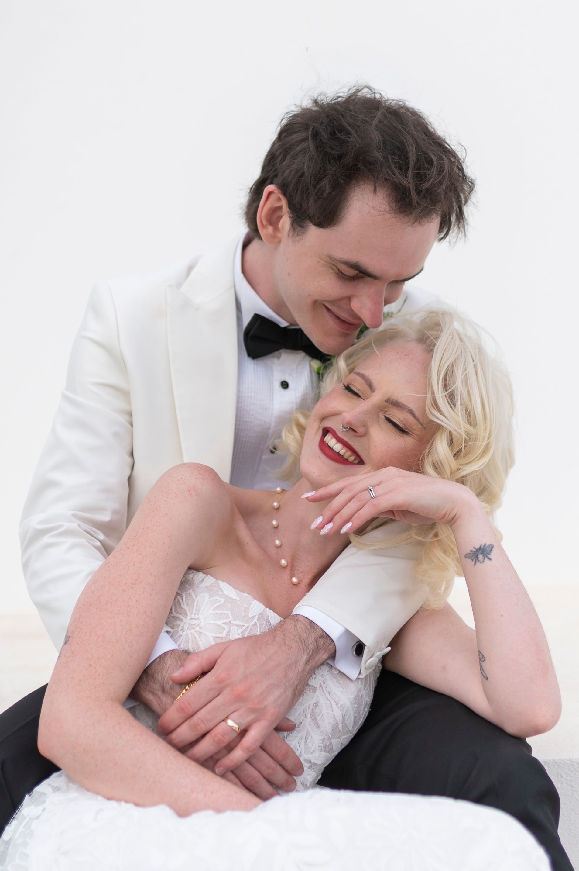 Bride and groom in wedding attire embrace and laugh together, groom in white tuxedo, bride in lace dress with pearl necklace.