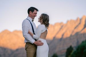 Couple embracing at sunset with dramatic mountain backdrop, wearing wedding attire in an outdoor romantic setting.