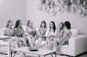 Seven women in matching robes sit together on a white sofa, laughing and relaxing, with straw hats displayed on the wall.