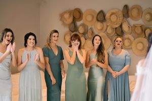 Six bridesmaids in sage green dresses react with joy and tears seeing the bride for the first time before the wedding ceremony.