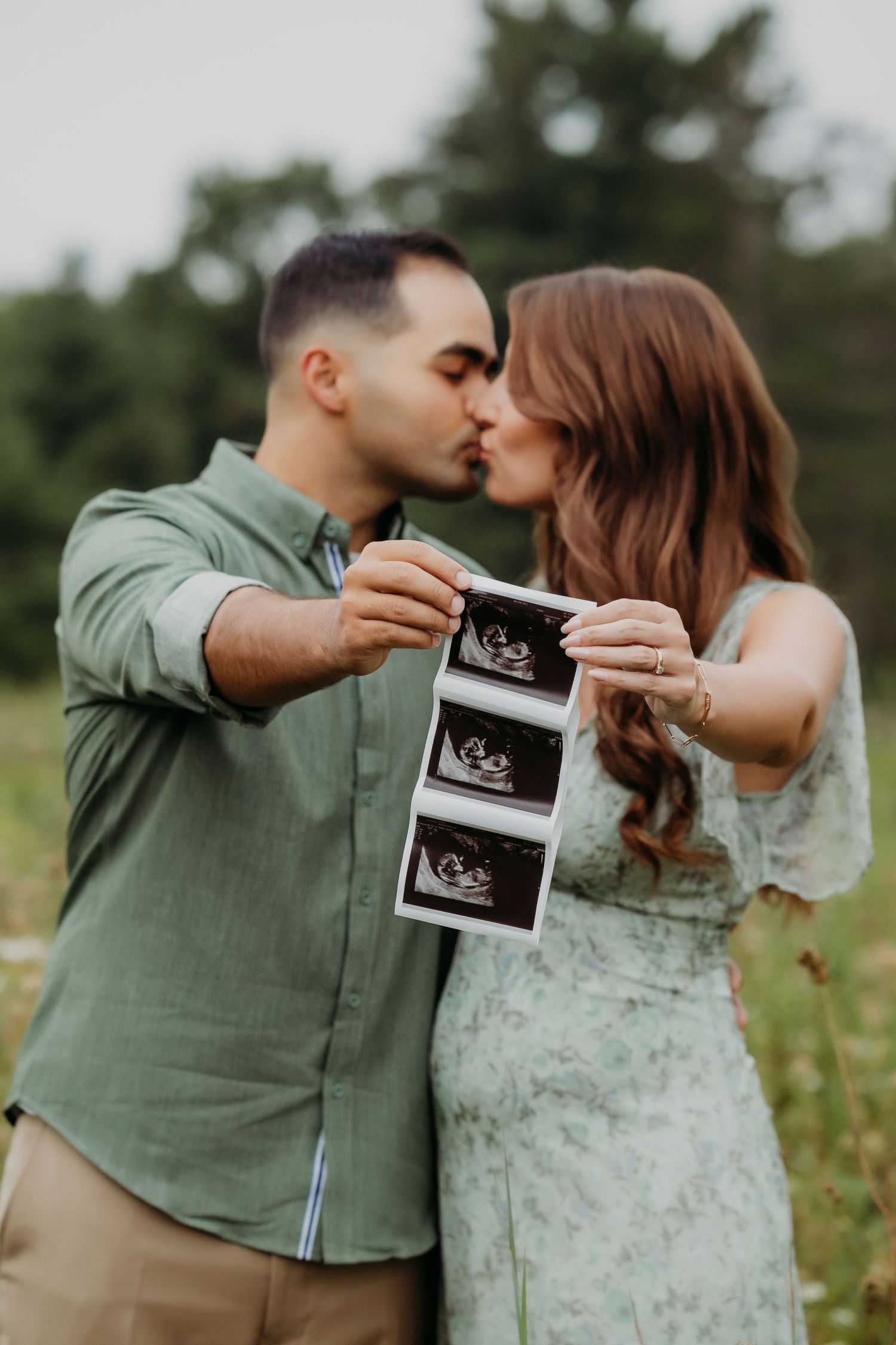 Couple kissing outdoors while holding ultrasound photos, announcing pregnancy in a green field setting.
