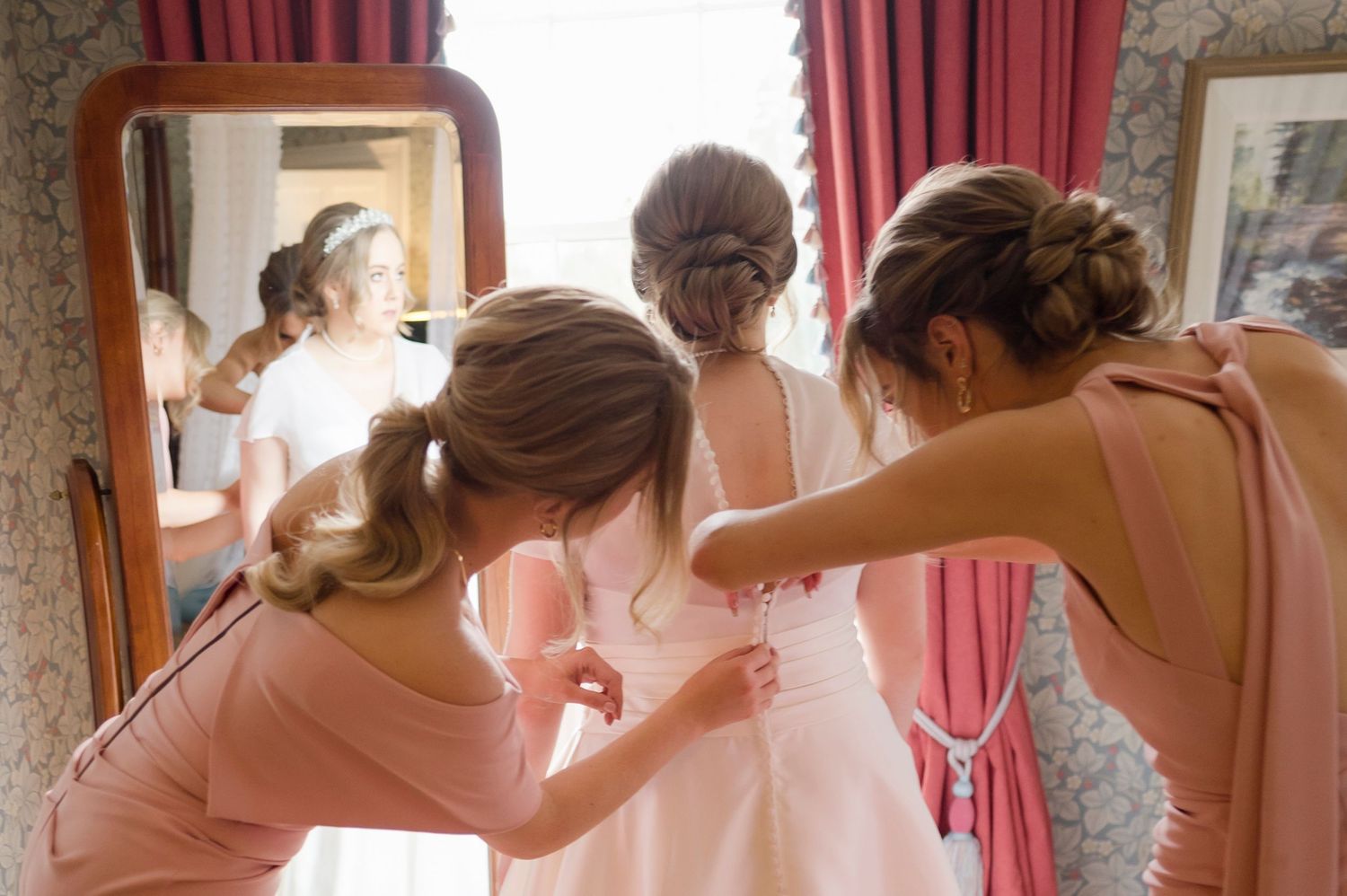 Two bridesmaids in pink dresses helping a bride with her white wedding gown in front of a mirror at Boyne Hill House, Co. Meath