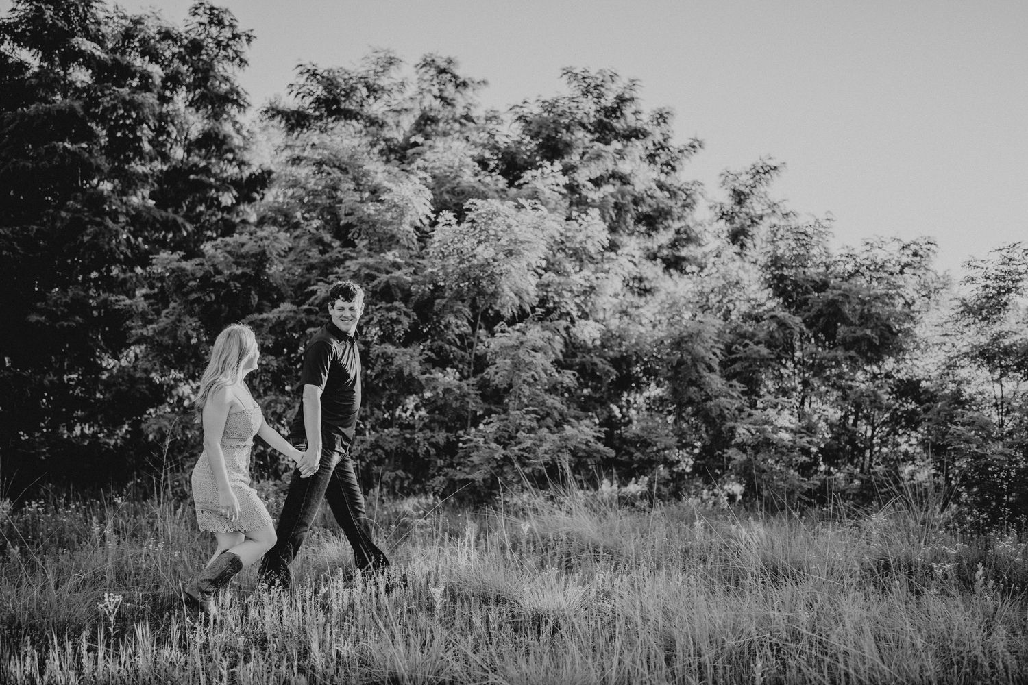 Black and white photo of a couple holding hands walking through a lush green meadow with trees in the background.