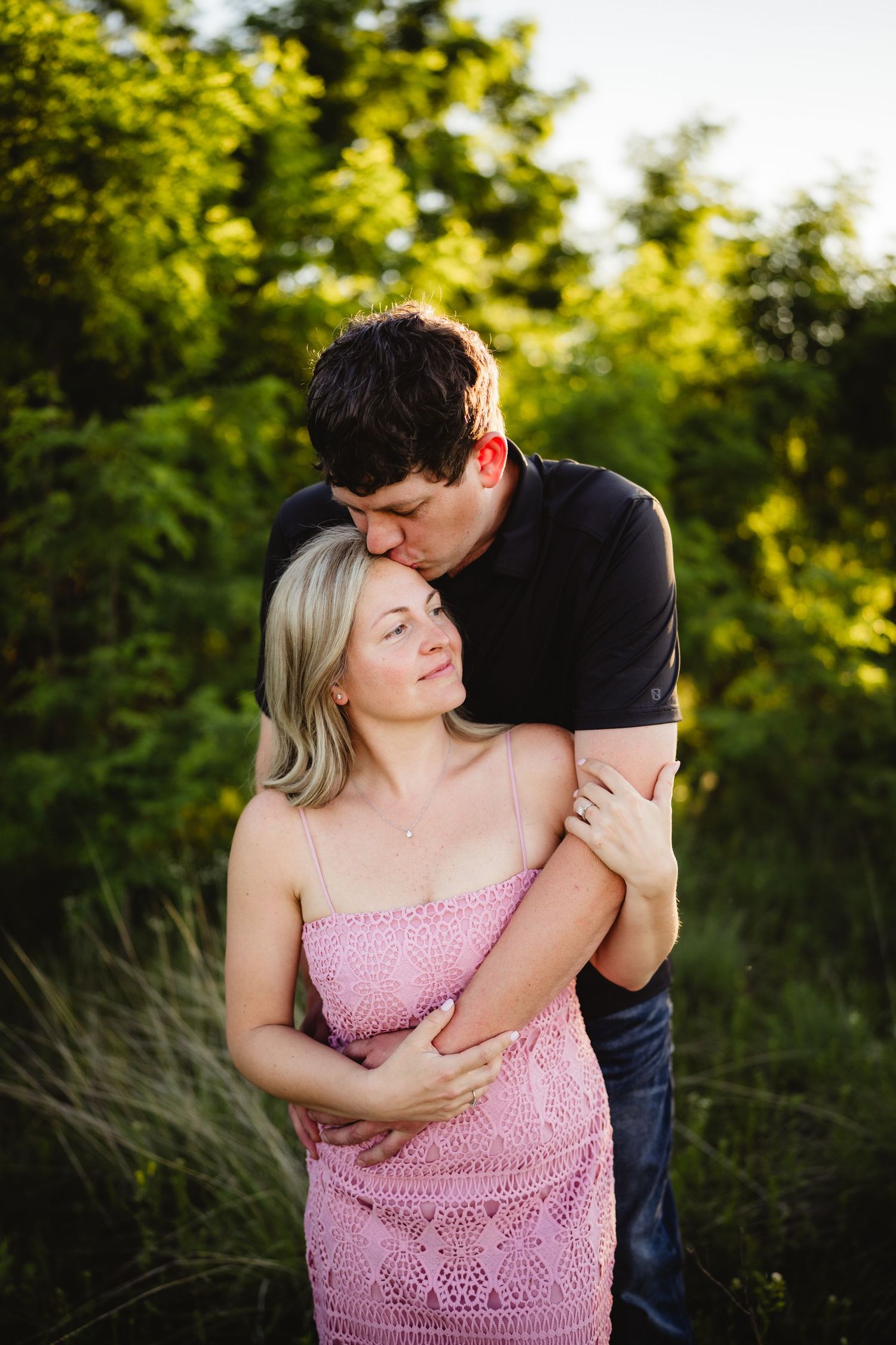 A couple embraces outdoors, man kissing woman's forehead, she wears pink dress, green trees in background.