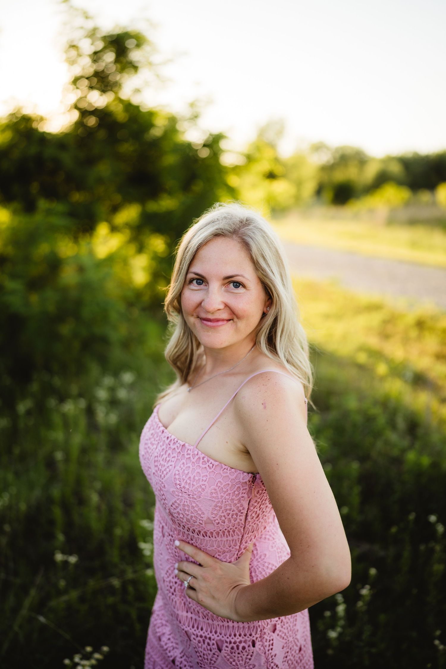 Smiling blonde woman in a pink lace sundress standing in a sunlit green field during golden hour.