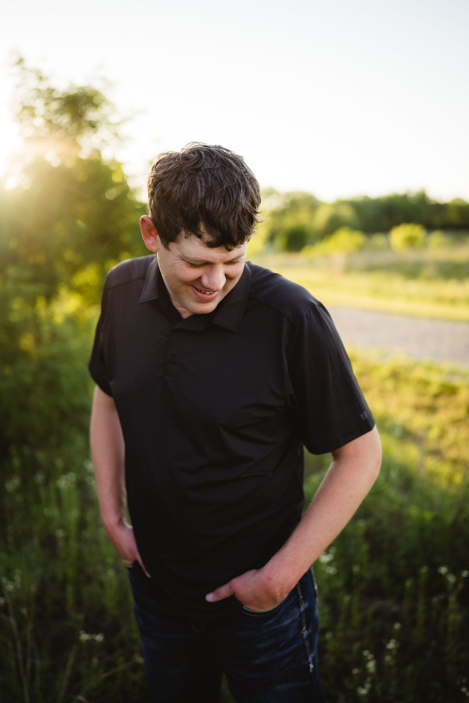 Young man in black polo shirt smiling outdoors at golden hour, standing in green field near road.