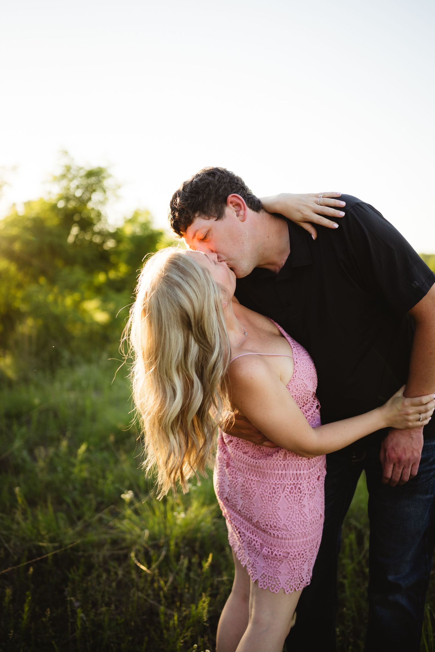 Couple sharing a romantic kiss outdoors at golden hour, woman in pink dress embracing man in black shirt.