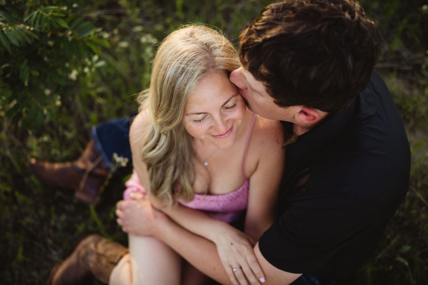 Couple sharing a tender moment outdoors, woman smiling as man kisses her cheek near lush greenery.