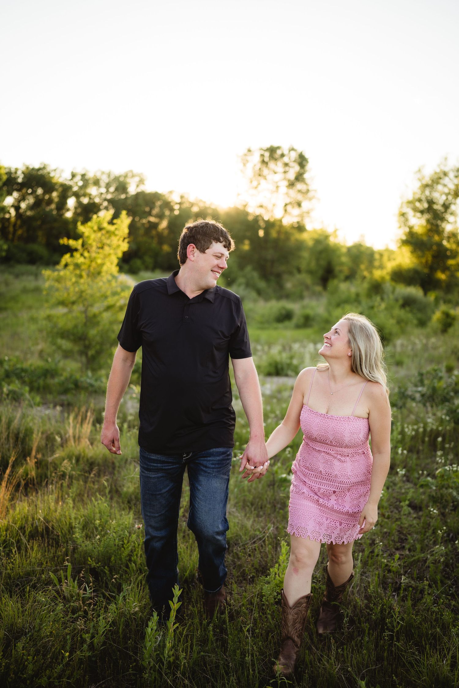 Couple holding hands walking through green field at golden hour, woman in pink dress and cowboy boots, man in black shirt.
