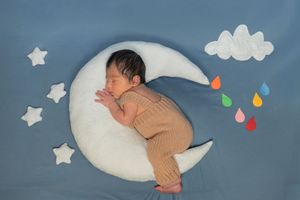 Newborn baby sleeping on a white crescent moon pillow with stars and colorful raindrops on a blue background.