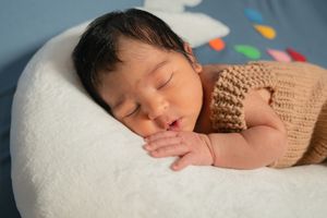 Newborn baby sleeping peacefully on white pillow wearing brown knit outfit with colorful decorations in background.