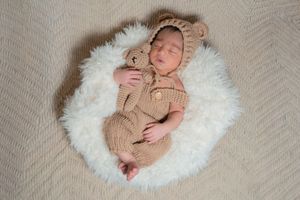Sleeping newborn baby in knit bear outfit and bonnet cuddling a teddy bear on white fluffy rug.
