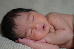 Newborn baby sleeping peacefully on an adult hand, with dark hair and rosy lips, captured in a serene close-up photo.