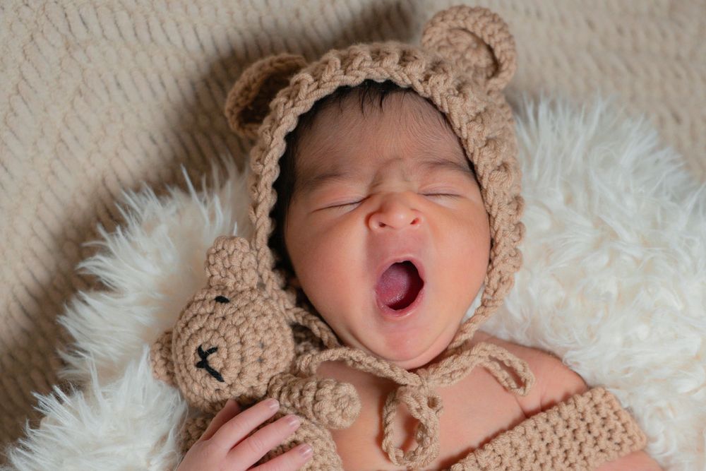 Adorable newborn baby yawning, wearing a cozy crocheted teddy bear bonnet and outfit, lying on a fluffy white blanket.