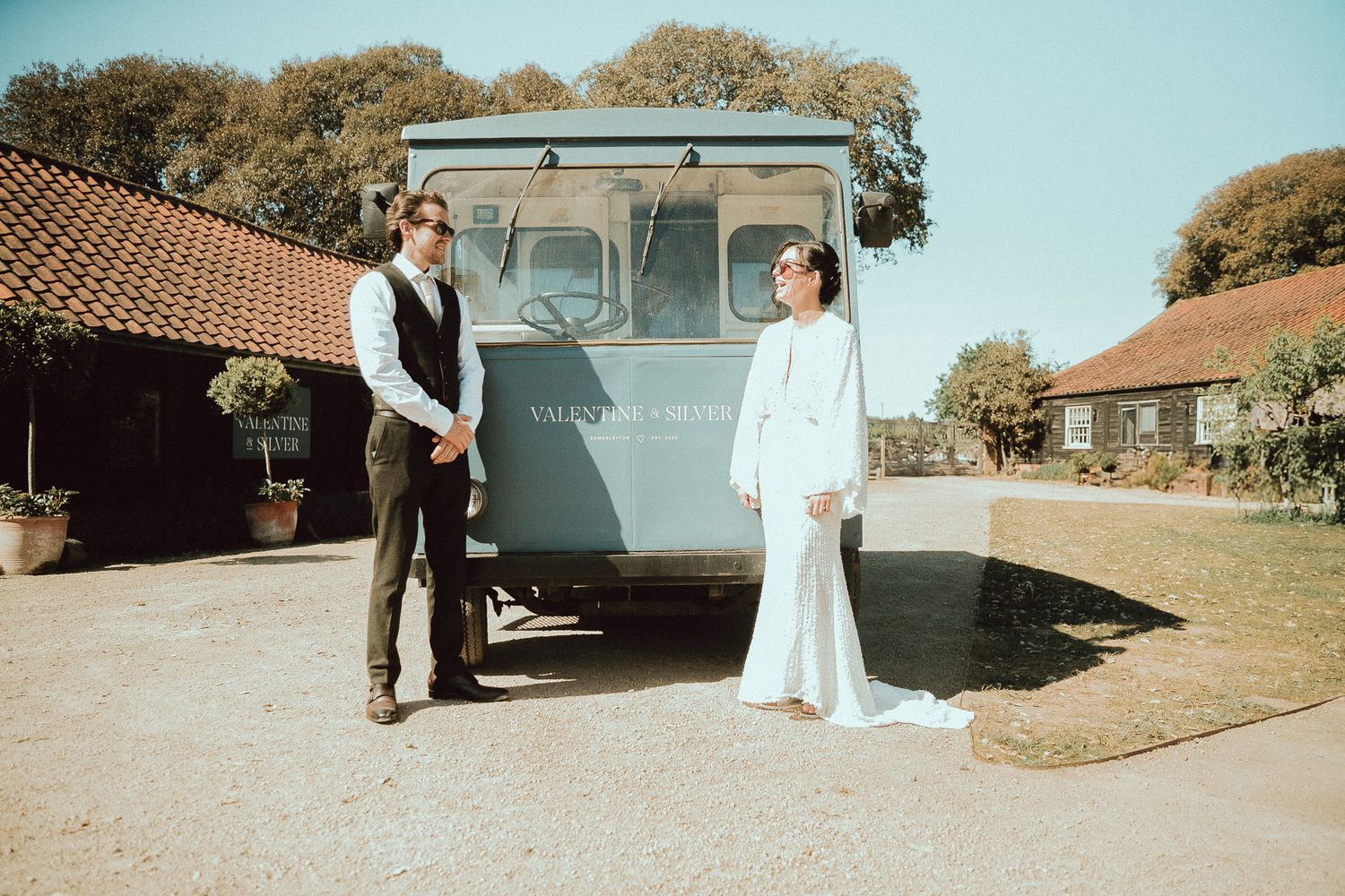 Bride and groom holding hands in front of a vintage blue van at a rustic farm wedding venue on a sunny day.