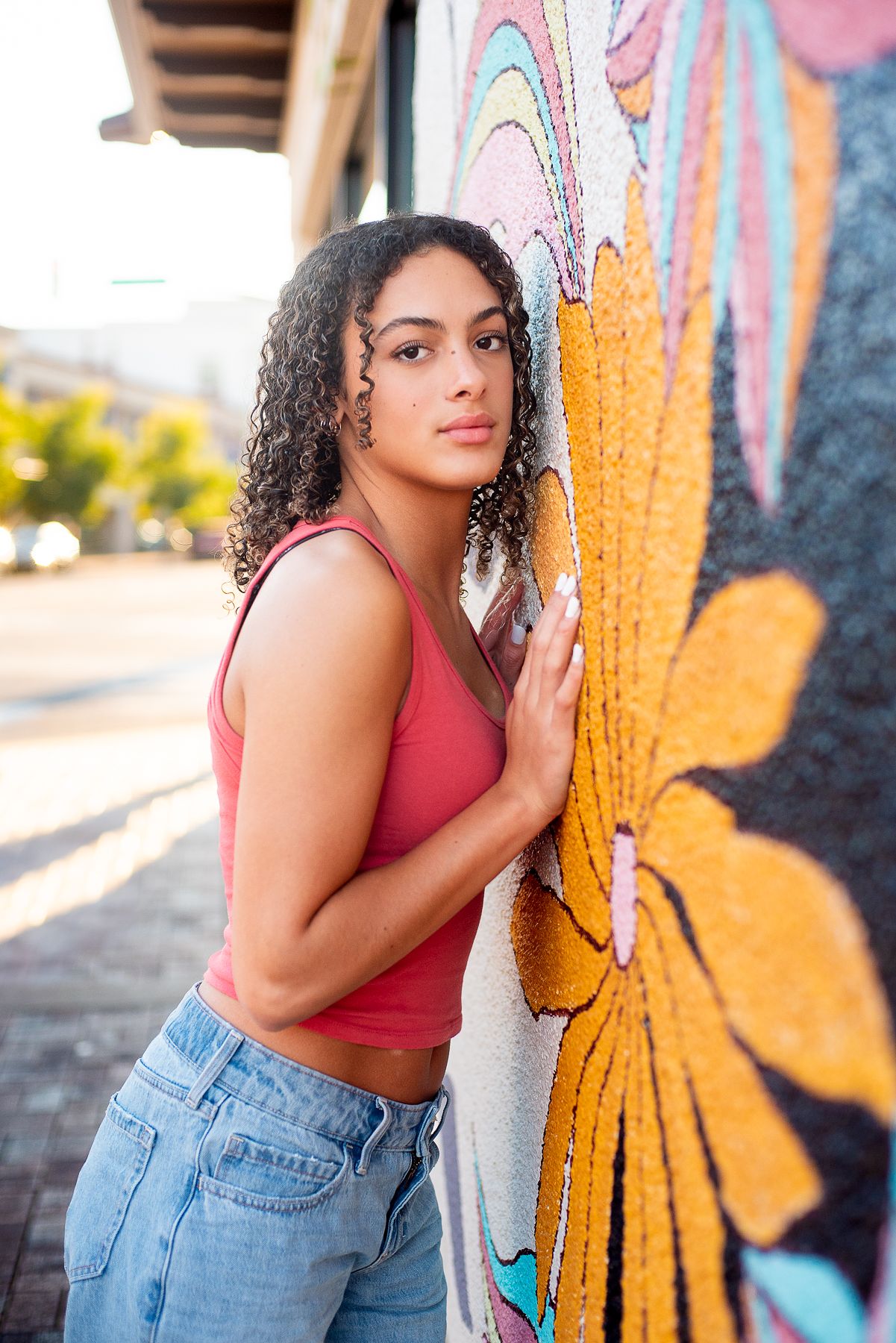 Young woman with curly hair in pink tank top and jeans leaning against colorful floral mural outdoors.