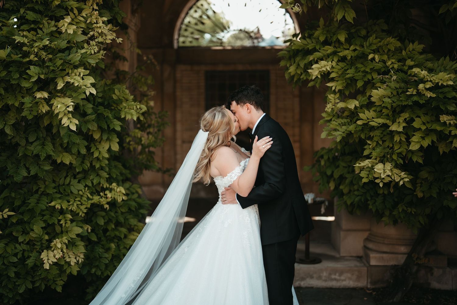 A bride and groom share a romantic kiss surrounded by lush greenery at an elegant outdoor wedding venue.
