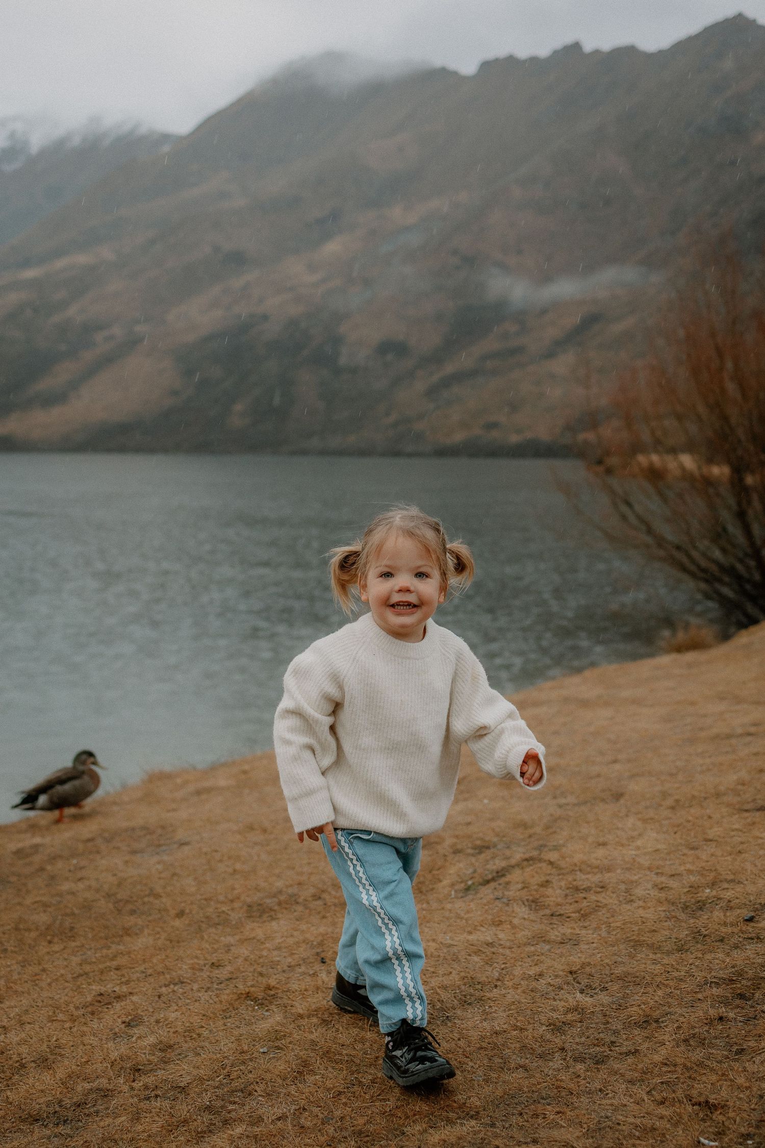 A happy toddler girl in a white sweater and blue pants runs near a scenic mountain lake on a cloudy day.