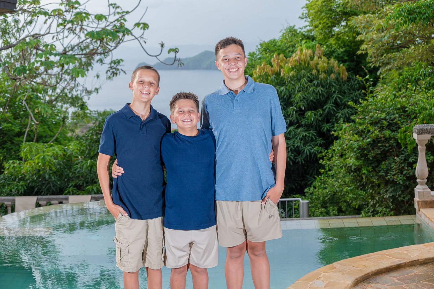 Three men in blue polo shirts and khaki shorts smiling together by a tropical pool with lush greenery.