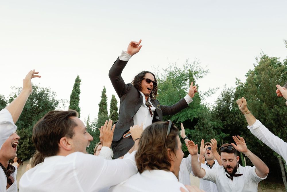 Groom in sunglasses lifted by groomsmen in white shirts outdoors at wedding celebration.