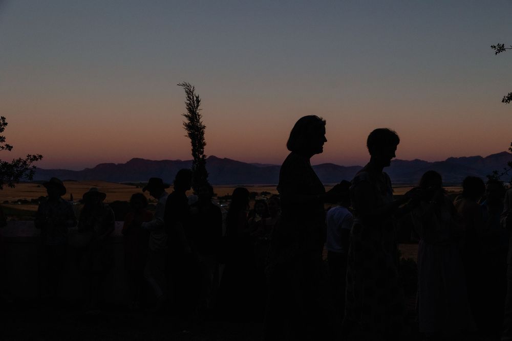 Silhouettes of people gathered outdoors against a vibrant sunset sky with mountain ridges in the background.