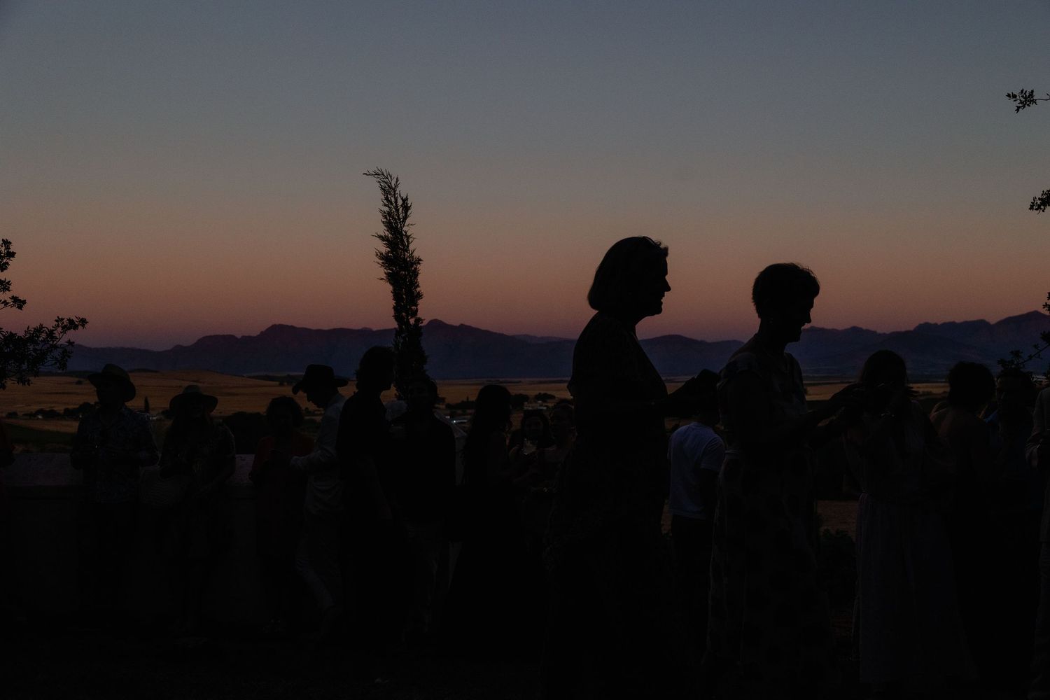 Silhouettes of people gathered outdoors against a vibrant sunset sky with mountain ridges in the background.