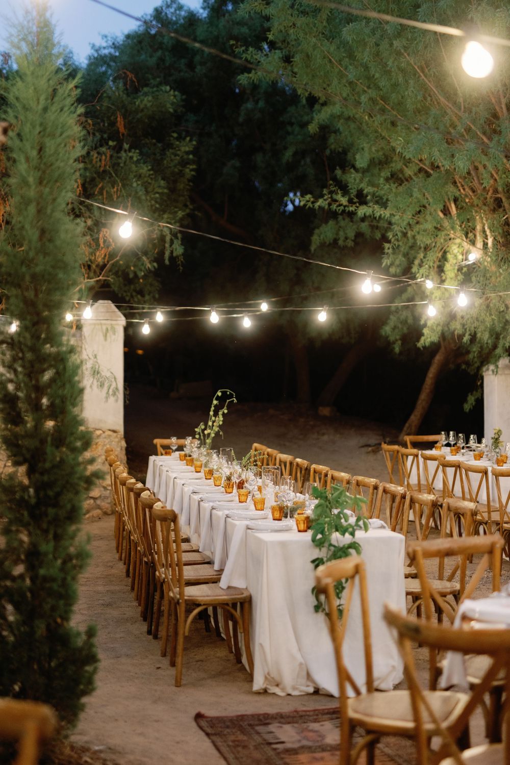 Elegant outdoor wedding reception with long white-draped tables, wooden cross-back chairs, and warm string lights hanging above lush trees.