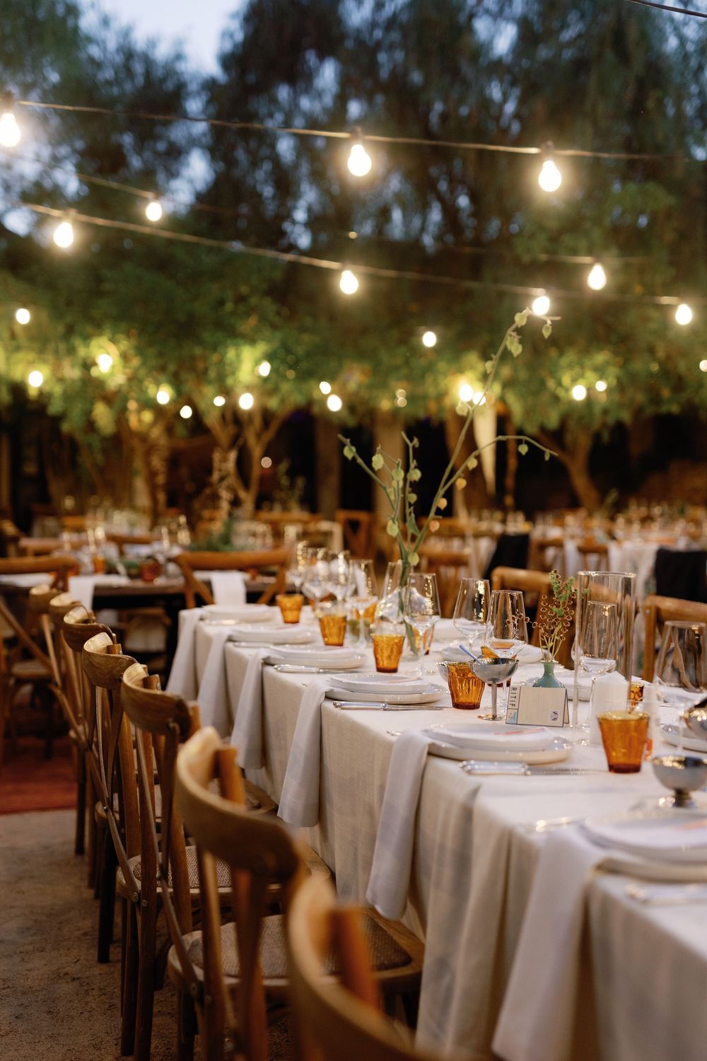 Elegant outdoor wedding reception with string lights, white tablecloths, amber glassware, and floral centerpieces at dusk.