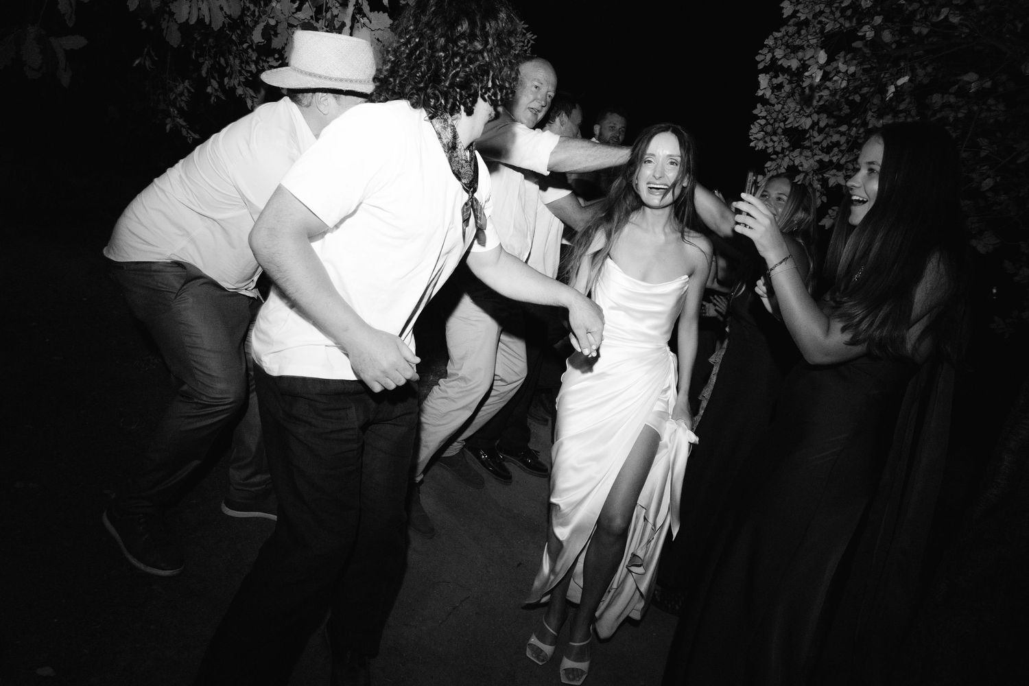 Black and white photo of guests dancing joyfully at an outdoor wedding reception at night.
