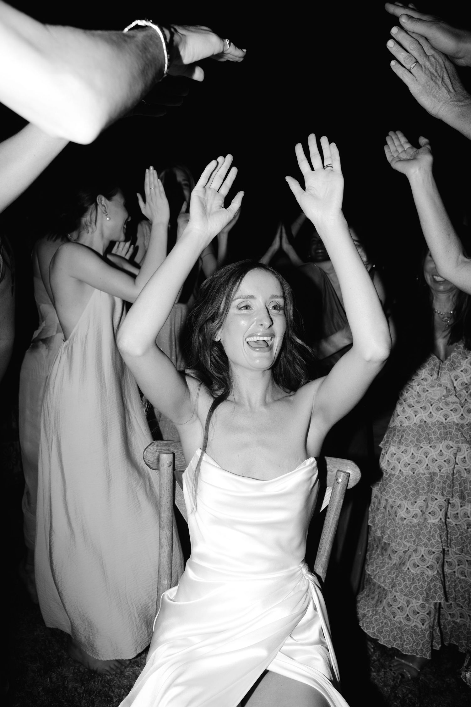 Bride sits in chair with hands raised, surrounded by dancing wedding guests in black and white photo.