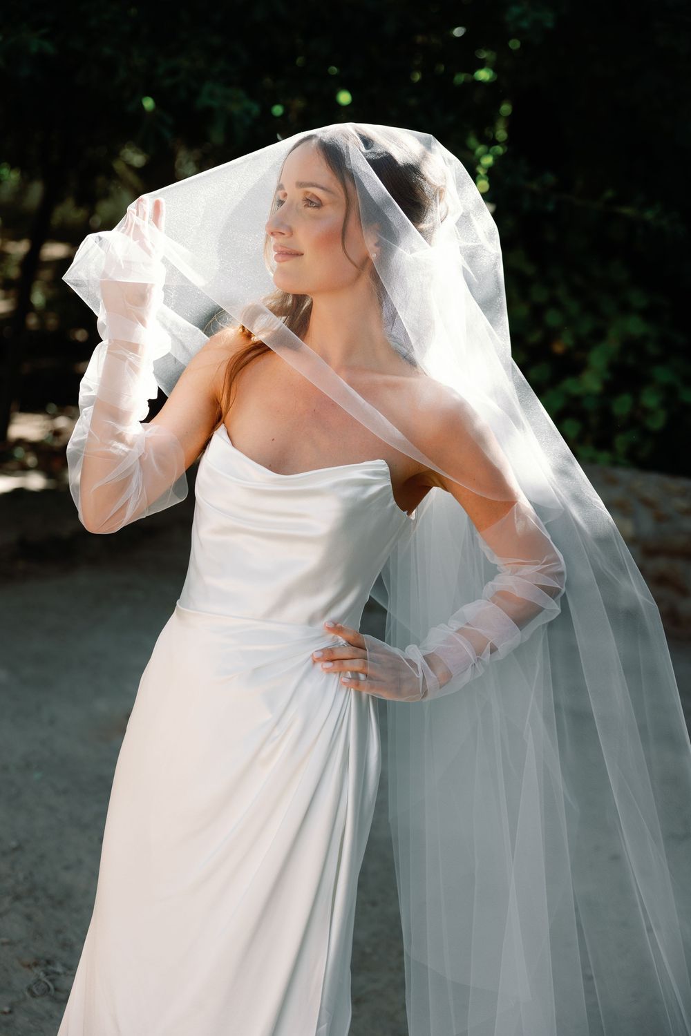 Elegant bride in strapless white gown and sheer veil with tulle gloves posing outdoors in sunlight.