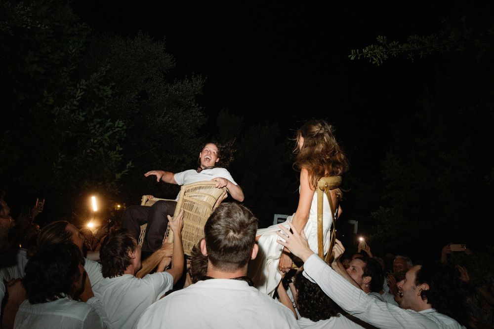 Bride and groom lifted by guests during lively outdoor wedding celebration at night.