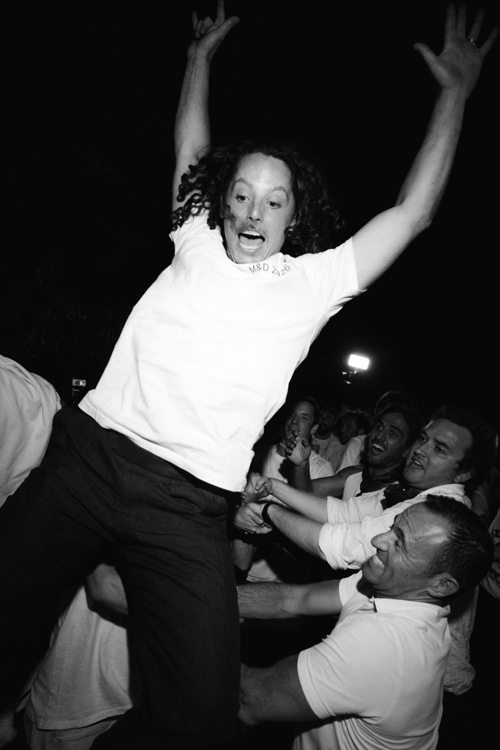 Woman with curly hair raises arms joyfully while being lifted by crowd at outdoor nighttime celebration.