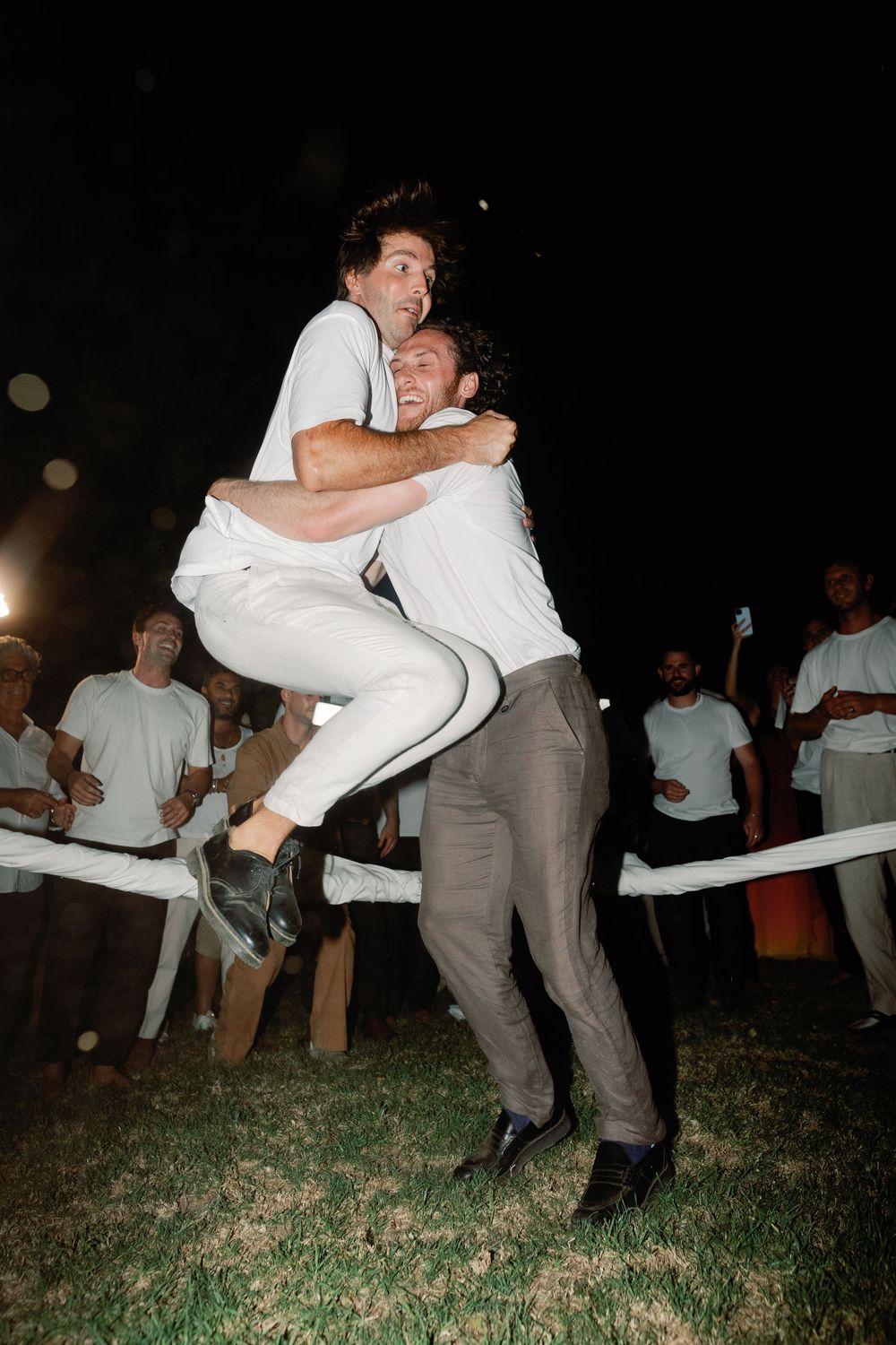 Two men in white embrace as one lifts the other at an outdoor nighttime wedding celebration.