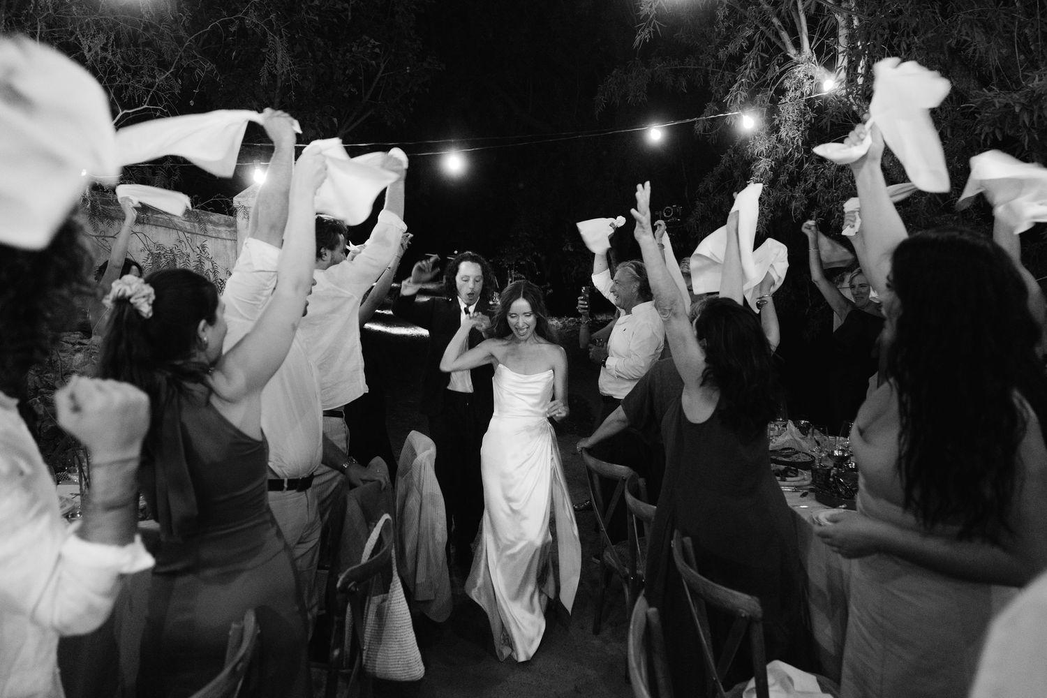 Black and white photo of bride and groom walking through guests waving white napkins at outdoor wedding reception.