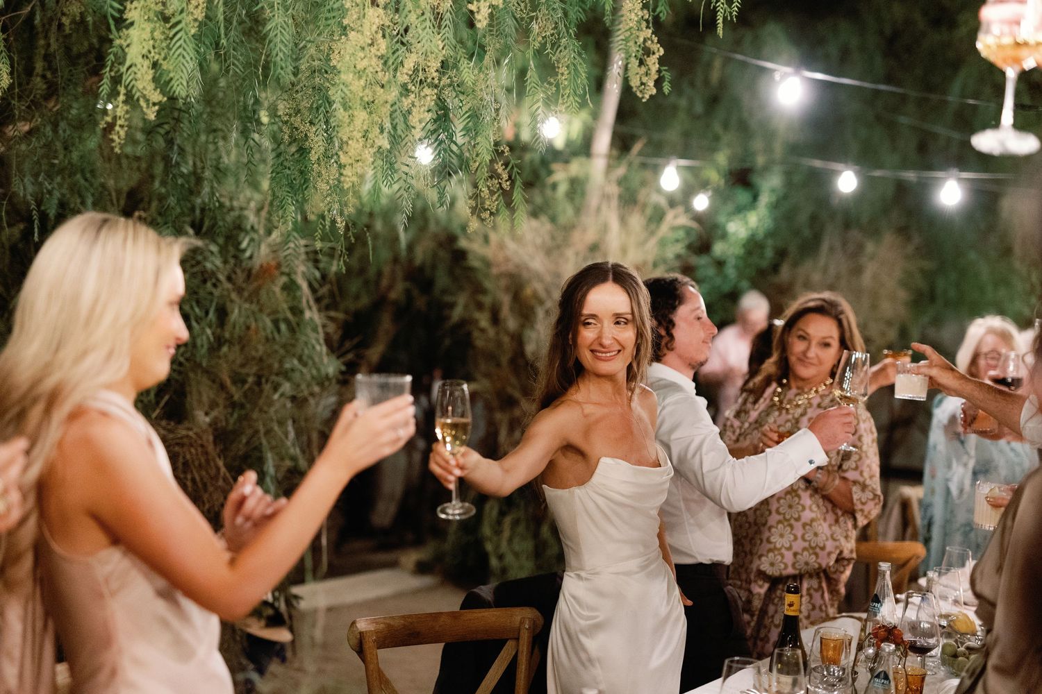 Bride in white gown toasting champagne with guests at elegant outdoor wedding reception with string lights.