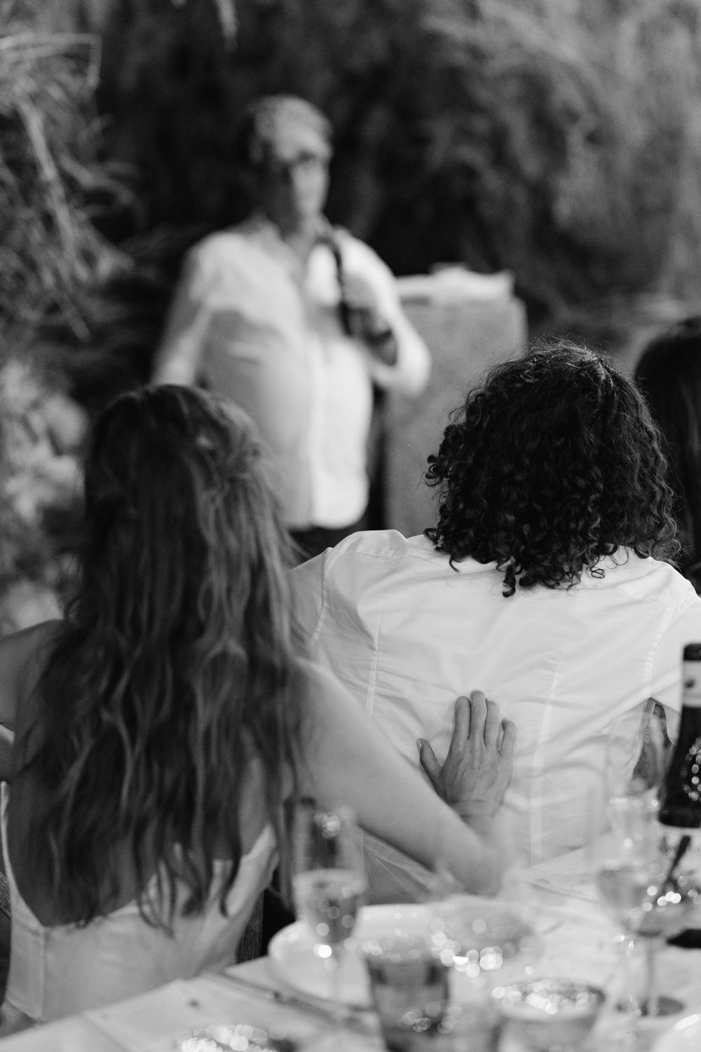 Black and white photo of wedding guests seated at a table listening to a speech during an outdoor reception.