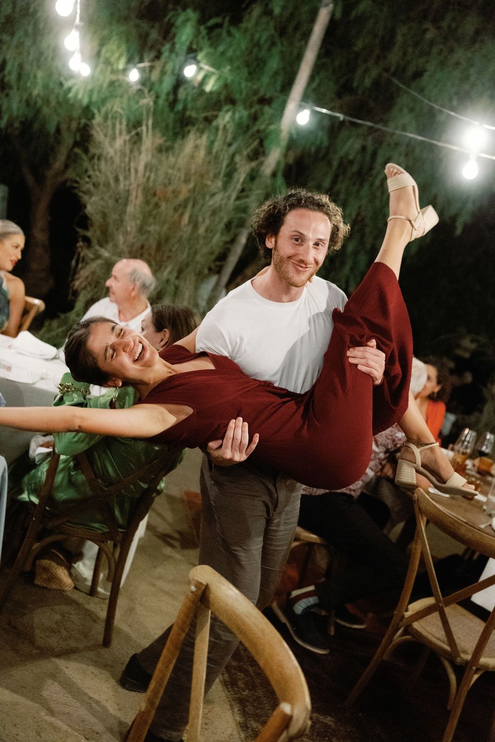 A man lifts a smiling woman in a burgundy outfit at an outdoor evening wedding reception with string lights.
