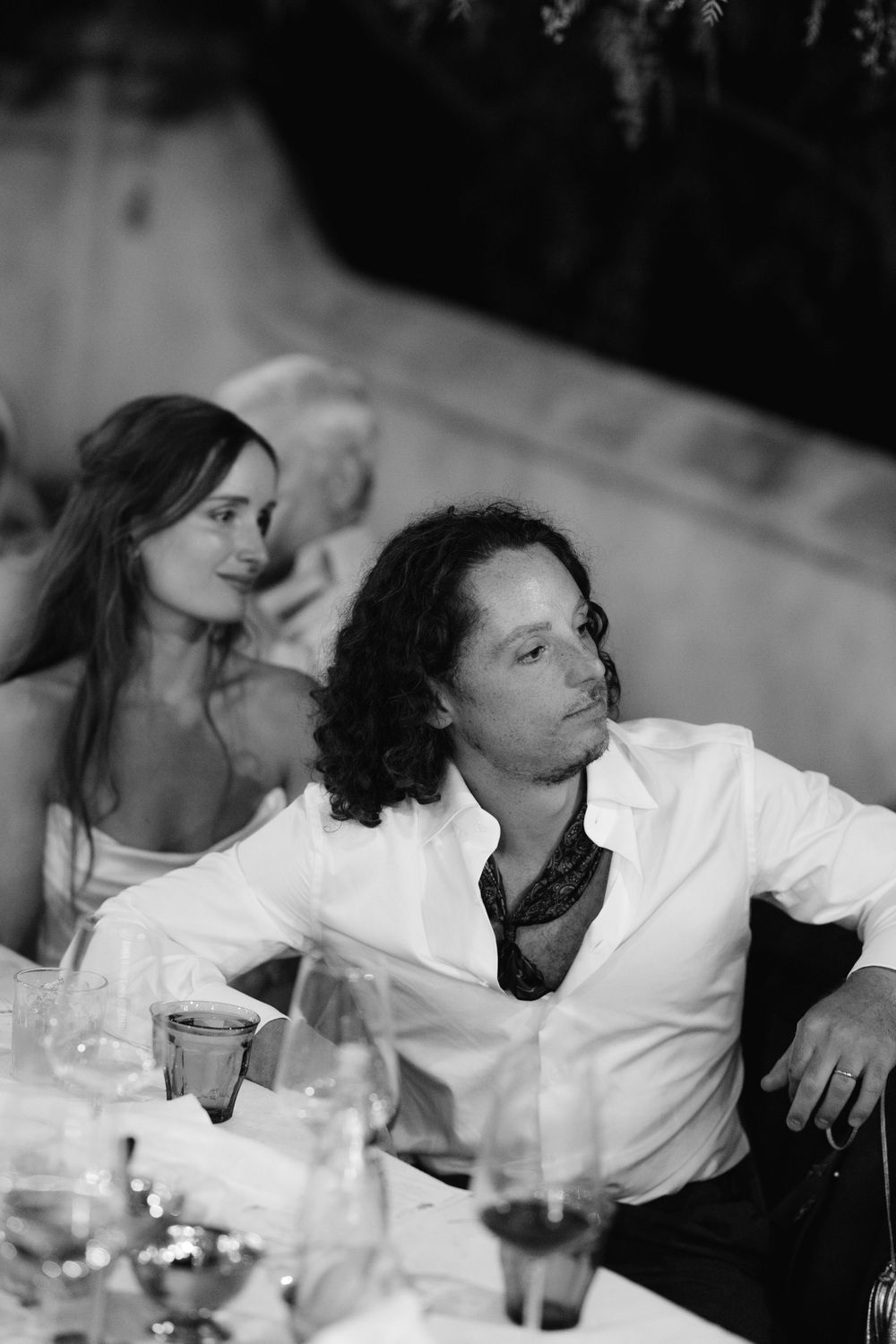Black and white photo of a couple seated at a wedding reception table, man in white shirt relaxing.