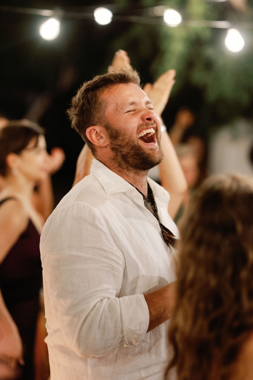 A bearded man in a white linen shirt laughs joyfully at an outdoor evening party with string lights.