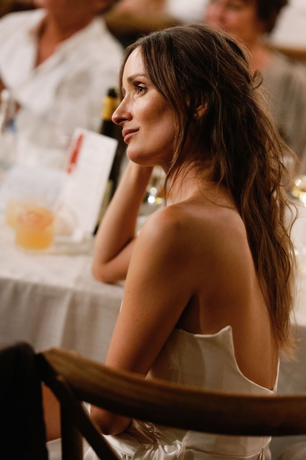 Elegant bride in a strapless white gown sits thoughtfully at a warmly lit wedding reception table.