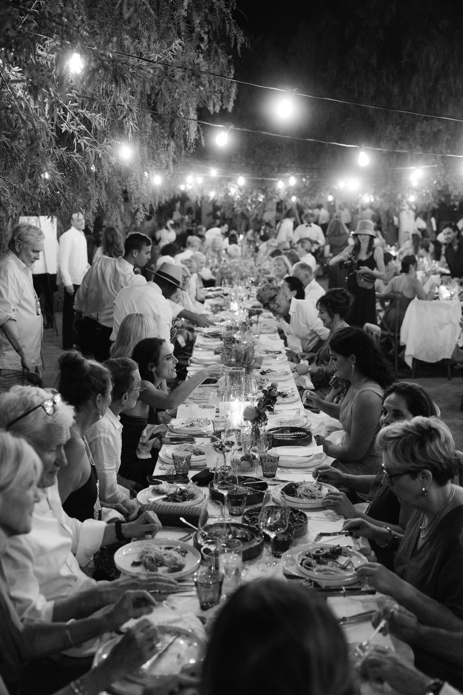 Guests dine at long outdoor wedding reception table with string lights overhead in black and white.