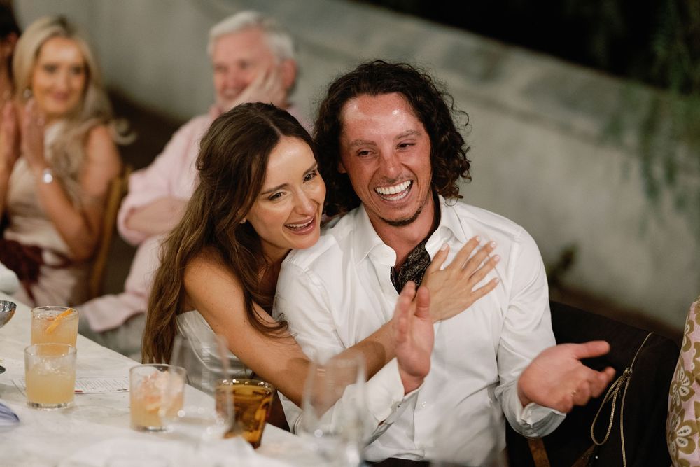 Couple smiling and laughing together at a wedding reception dinner table, bride in white dress hugging groom.
