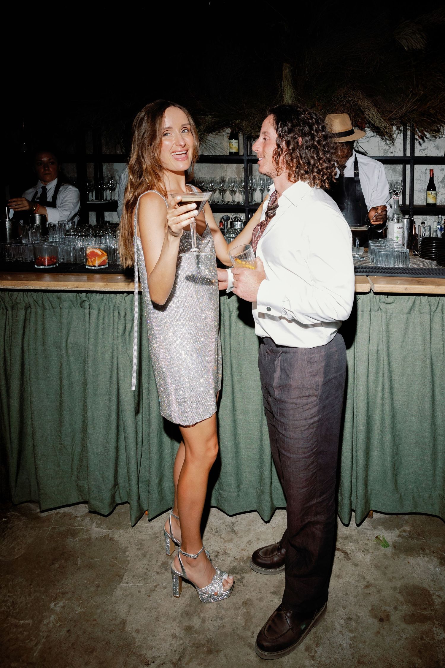 Couple laughing and holding cocktails at a bar during an elegant party, woman in silver sequin dress and heels.