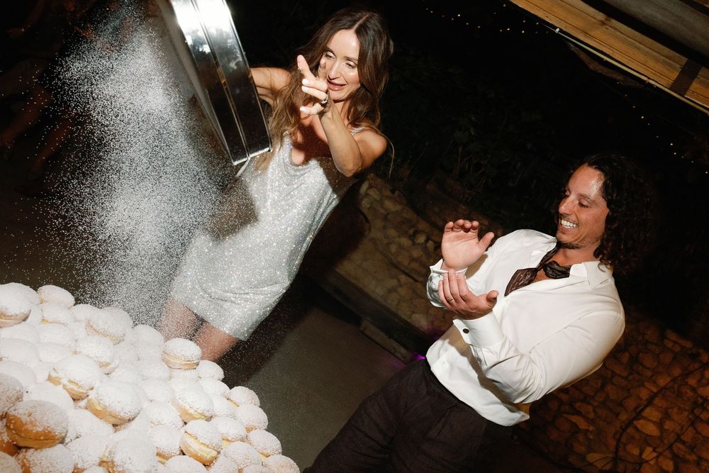 Bride and guest playfully toss powdered donuts at nighttime wedding reception dessert station.