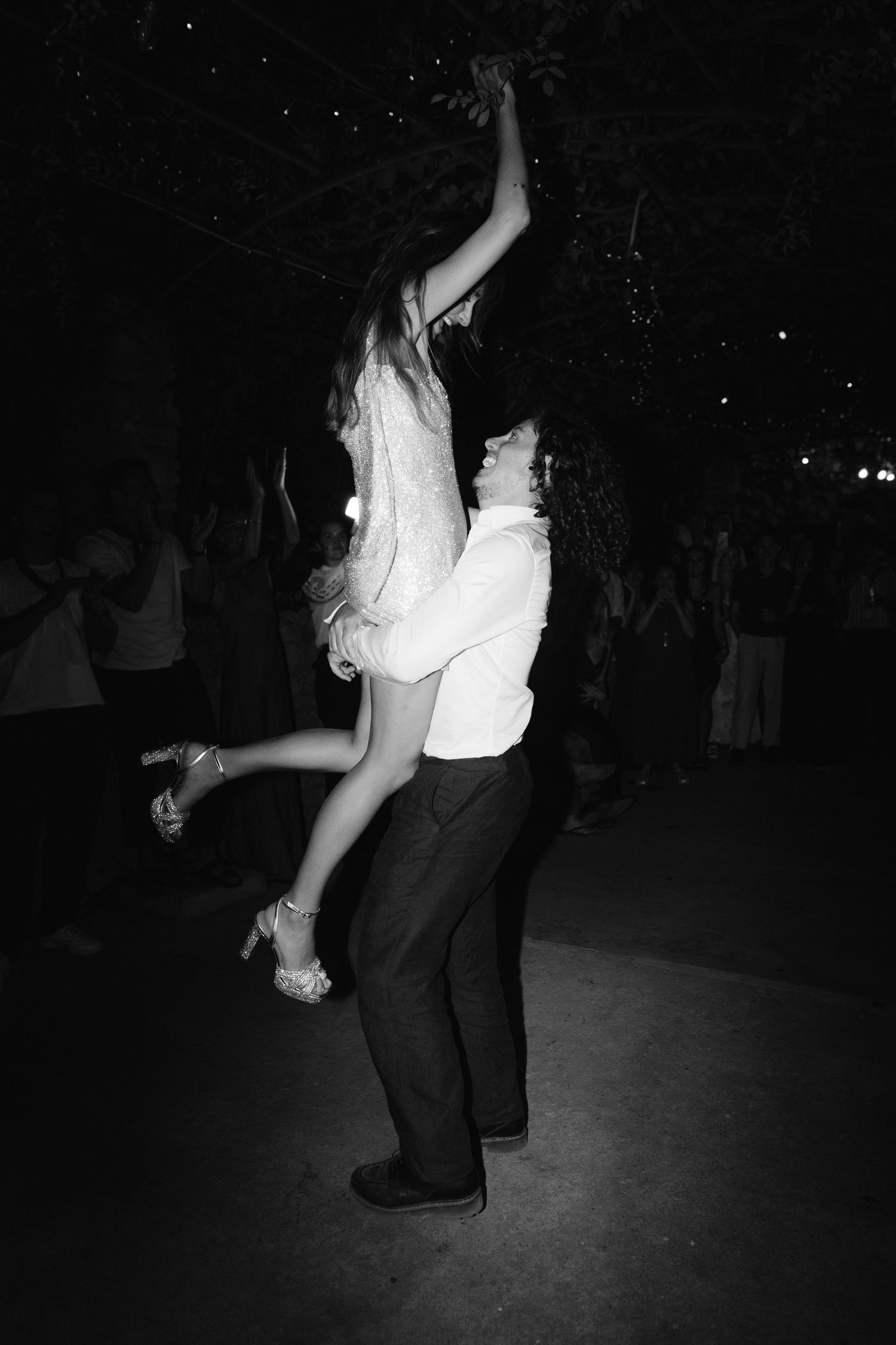 Bride and groom dancing energetically outdoors at night, groom lifting bride in wedding attire.