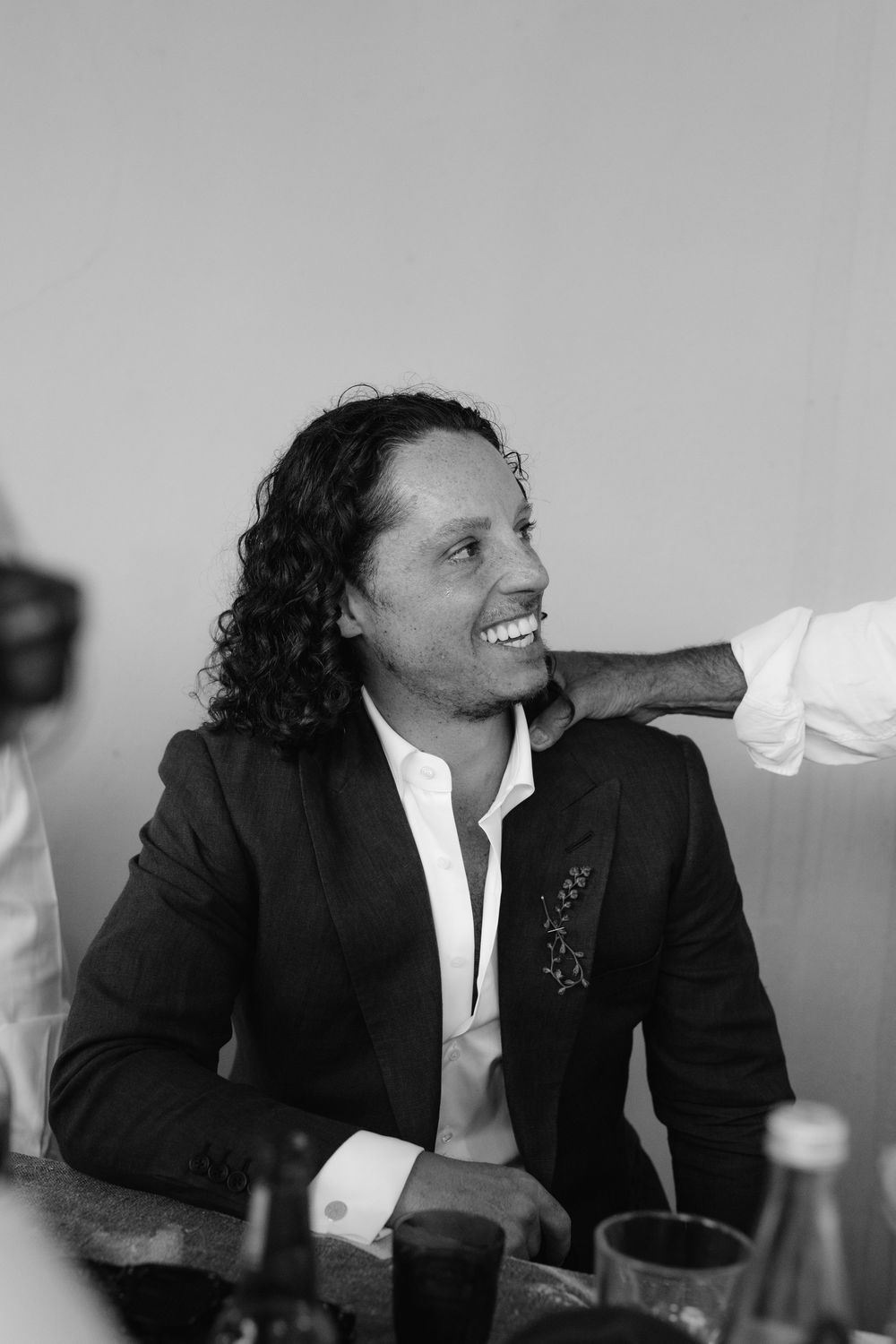 Smiling groom with curly hair in dark suit at wedding reception, black and white photo.